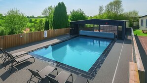 Ein rechteckiger Swimmingpool mit Holzterrasse und Liegestühlen, umgeben von einem Holzzaun und Grünpflanzen.