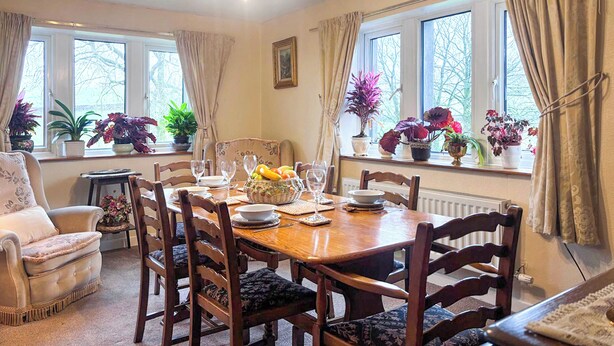 Comedor con una mesa de madera dispuesta para cuatro personas, rodeada de sillas. Hay plantas en macetas en el alféizar de la ventana y un candelabro cuelga del techo.