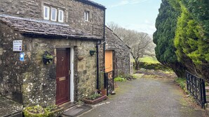 Una cabaña de piedra con puerta de madera, un pequeño jardín y un camino empedrado que lleva a una reja.