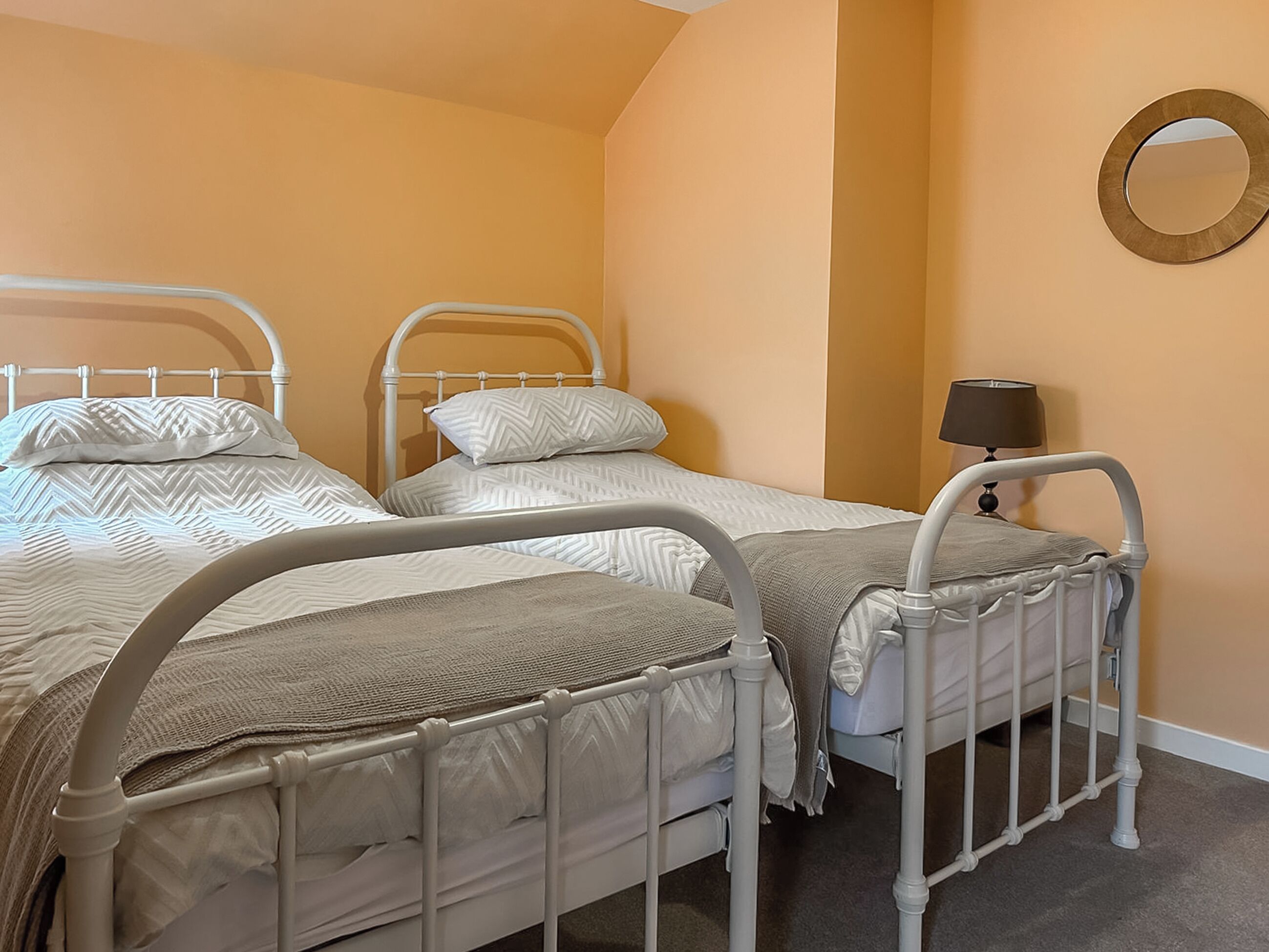 A room with three single beds, each with a white metal frame and beige bedding. There is a round mirror on the wall and a bedside table with a lamp.