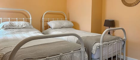A room with three single beds, each with a white metal frame and beige bedding. There is a round mirror on the wall and a bedside table with a lamp.