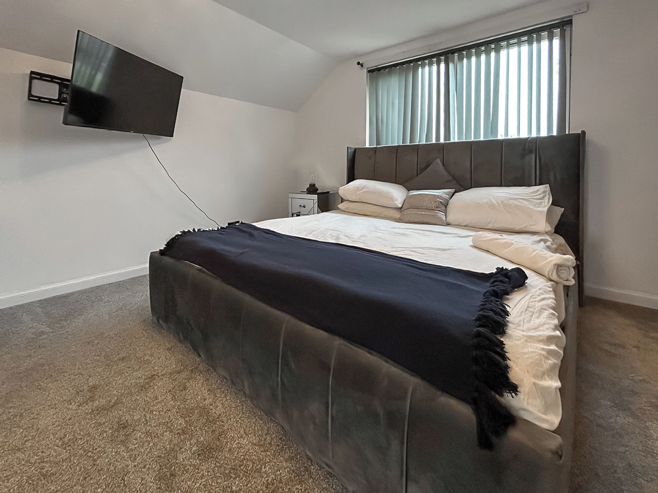 A bedroom with a bed, a television mounted on the wall, a nightstand, and a window with blinds.