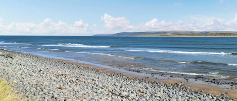 A pebbly beach with waves, a clear sky, and distant hills.