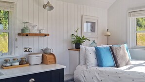 Un dormitorio ordenado con cama, mesita de noche y un taburete pequeño. Hay una ventana con persianas, un estante en la pared con frascos y una tetera, y un cuadro enmarcado sobre la cama.