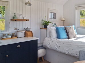 Un dormitorio ordenado con cama, mesita de noche y un taburete pequeño. Hay una ventana con persianas, un estante en la pared con frascos y una tetera, y un cuadro enmarcado sobre la cama.