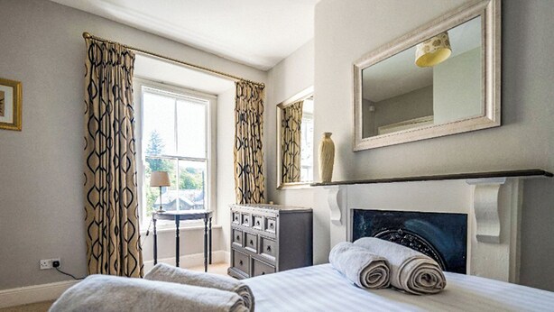 A hotel room with a bed, two rolled towels, a dresser, a mirror, and a window with curtains.