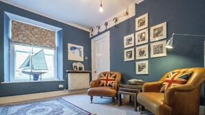 A room with a blue wall, a window with a sailboat decoration, two armchairs with Union Jack pillows, a patterned rug, and a framed picture collection on the wall.