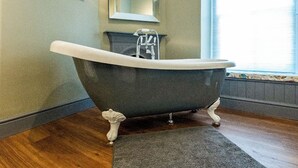 A clawfoot bathtub in a bathroom with a wooden floor, a mirror, and a window with blinds.