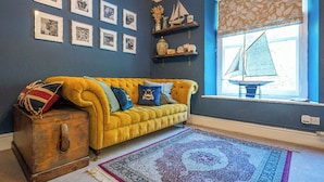 A room with a yellow sofa, a patterned rug, a wooden chest, and framed pictures on the wall.