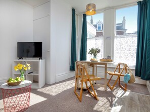 A modern living room with a television, a small table with a vase of flowers, a wooden chair, and a view of a building through large windows. - Bay Tree View - UK51428 (Paignton)