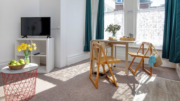 A modern living room with a television, a small table with a vase of flowers, a wooden chair, and a view of a building through large windows. - Bay Tree View - UK51428 (Paignton)