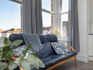 A dark leather sofa with floral cushions, a wooden side table, and a potted plant in a room with large windows and a view of buildings.