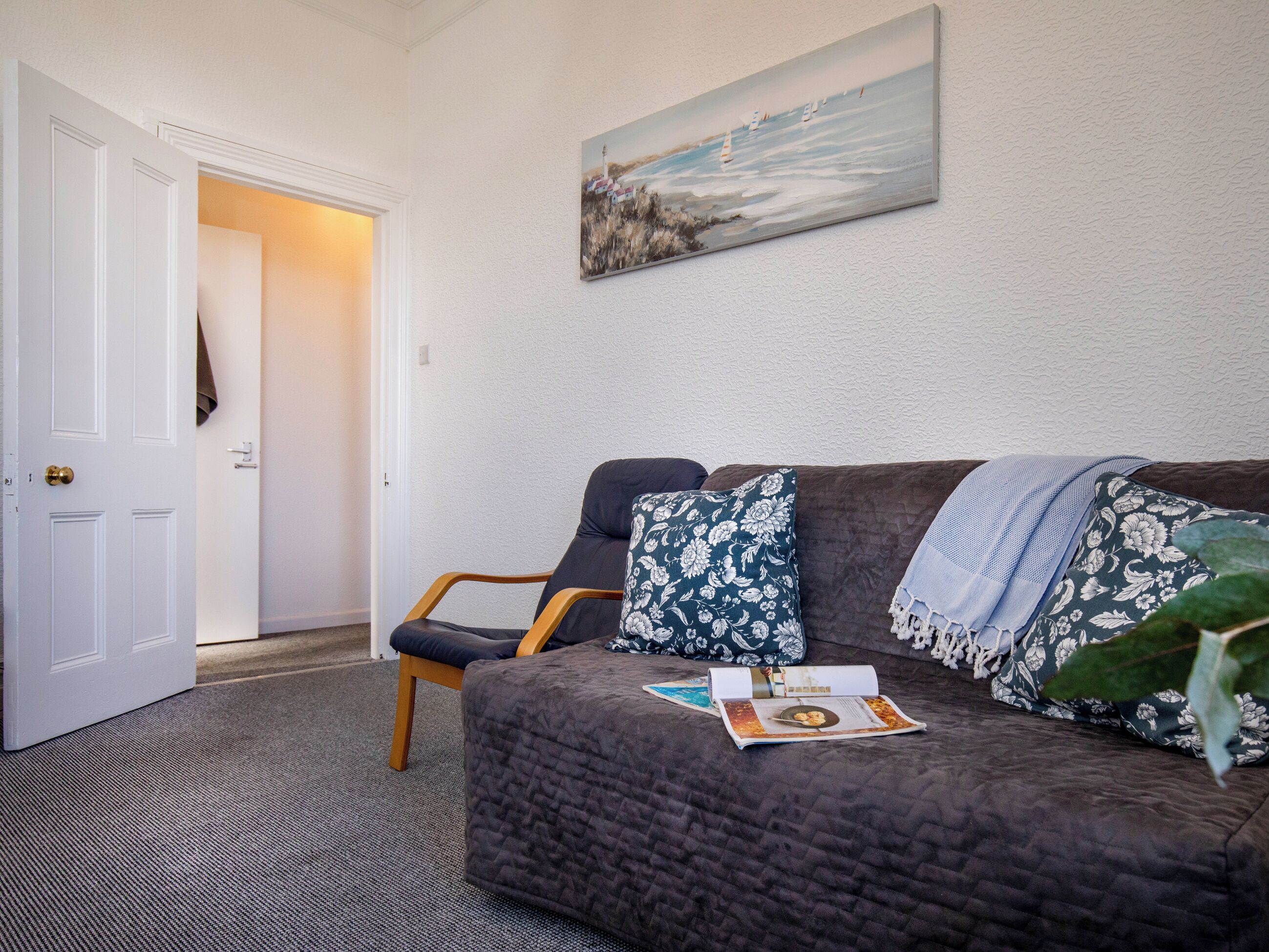 Ein Zimmer mit einem dunkelbraunen gesteppten Sofa, einem Holzstuhl, einer Tür und einem Gemälde einer Küstenlandschaft.