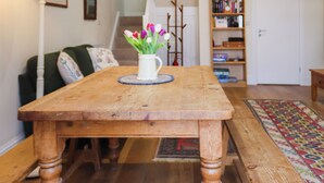 Een houten eettafel met een witte vaas vol bloemen, een groene bank, een boekenkast en een deur in een kamer met houten vloeren en een kamerbreed tapijt.