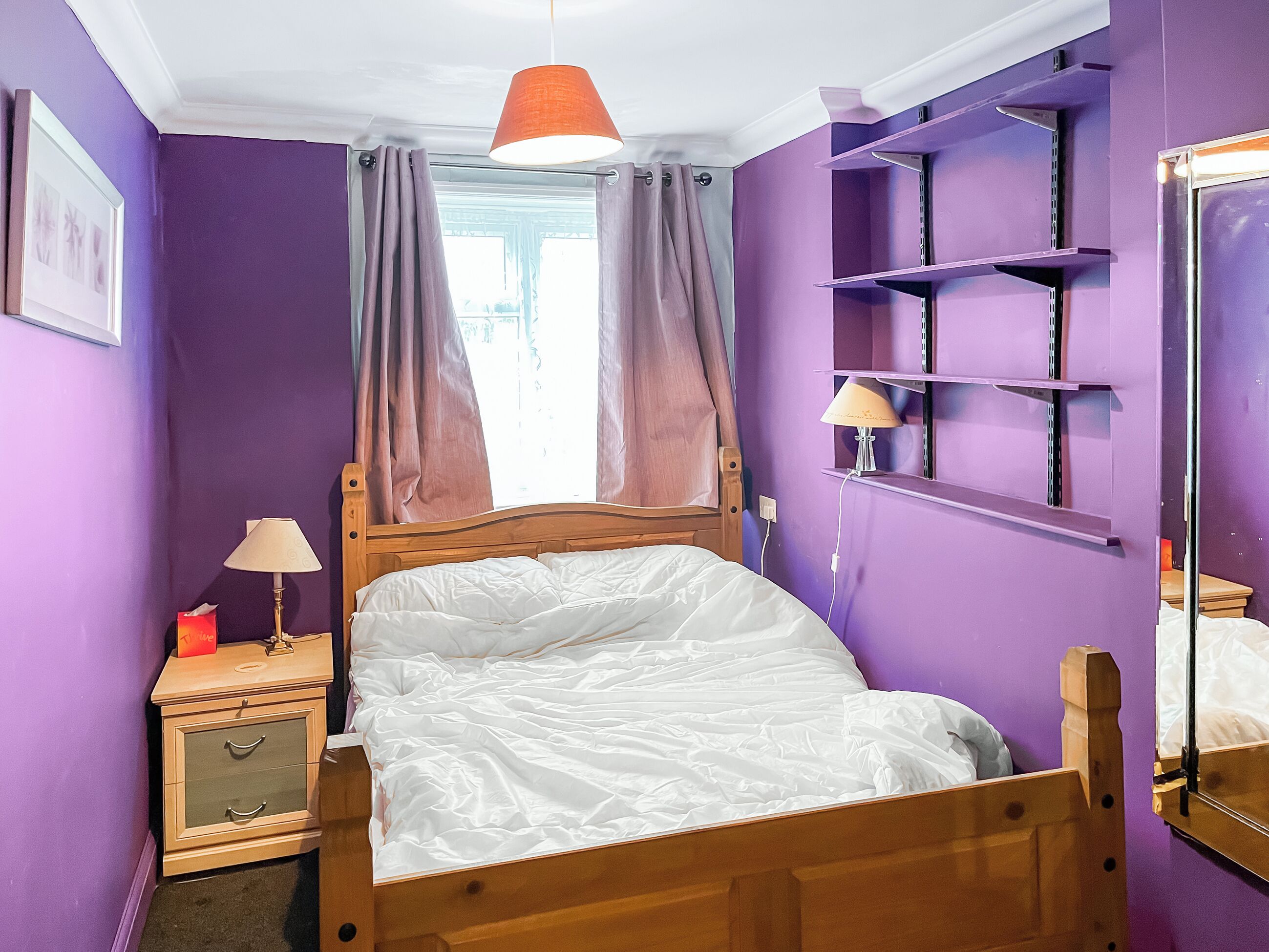 Een slaapkamer met een houten bed, paarse wanden, een raam met gordijnen, een nachtkastje met een lamp en een spiegel.