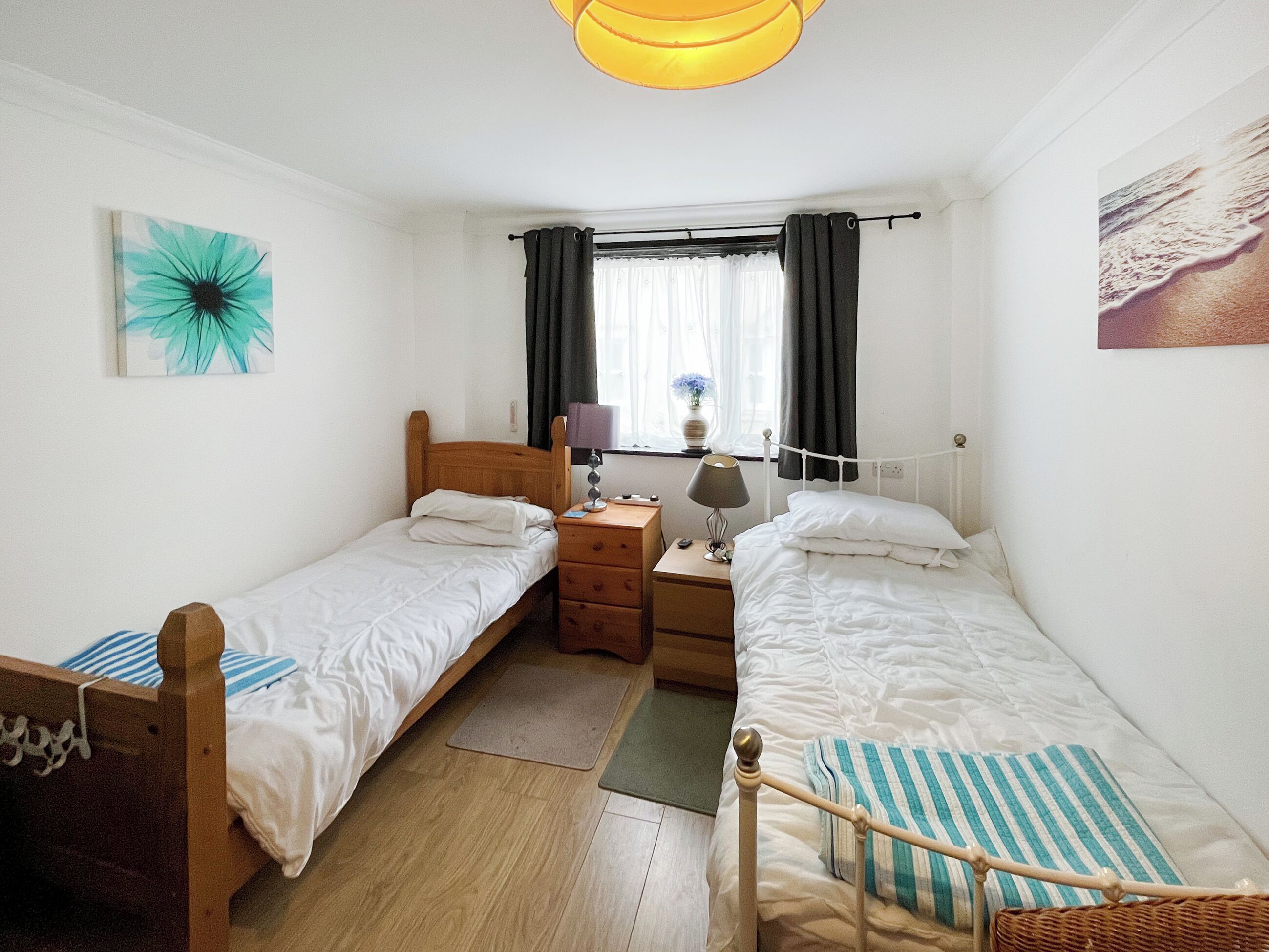 Una habitación con dos camas individuales, una mesita de noche de madera, un escritorio con lámpara, una ventana con cortinas y un cuadro en la pared.