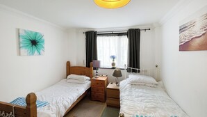 A room with two single beds, a wooden nightstand, a desk with a lamp, a window with curtains, and a wall painting. - 3 bedroom accommodation in Ramsgate (Ramsgate)