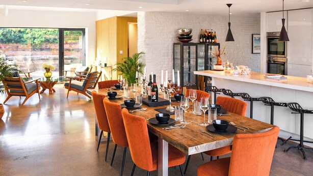 A modern dining area with a wooden table, orange chairs, and a kitchen in the background. - 5 bedroom accommodation in Heydon (Heydon)