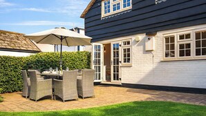 A patio with wicker furniture and a table, a hedge, and a house with a dark exterior and white walls. - Welcoming larger family gatherings. (Winterton-on-Sea)