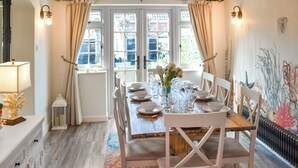 A dining room with a wooden table set for six, white chairs, a vase with flowers, and a colorful rug. - Welcoming larger family gatherings. (Winterton-on-Sea)
