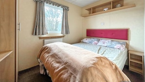 A bedroom with a bed, a window with curtains, a radiator, a shelf with items, and a light fixture.