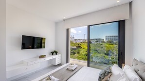 Un dormitorio moderno con una cama grande, un televisor montado en la pared, un aparador blanco y vistas a zonas verdes y edificios a través de amplias ventanas.