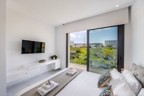 A modern bedroom with a large bed, a TV mounted on the wall, a white dresser, and a view of greenery and buildings through large windows. - Protaras By the Bay Villa 19 (Paralimni)