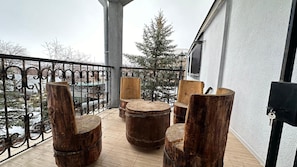 Un balcón con muebles de madera rústica, entre ellos sillas y una mesa redonda, con vistas a un paisaje nevado con árboles desnudos y un edificio al fondo.