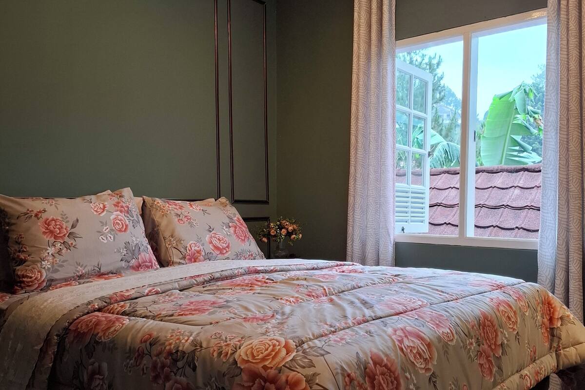 A bedroom with a floral bedspread, a nightstand with a vase of flowers, and a window with curtains.