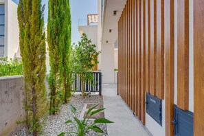A modern building with vertical wooden panels, a concrete wall, and a pathway with small trees and pebbles. - Protaras By the Bay Villa 19 (Paralimni)