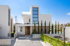 A modern building with a large glass window, a wooden door, and a concrete bench area. - Protaras By the Bay Villa 19 (Paralimni)
