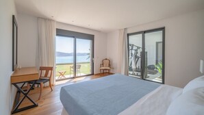 Ein Schlafzimmer mit einem großen Bett, einem Schreibtisch und einem Stuhl. Eine Glastür führt auf einen Balkon mit Meerblick.