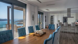 A modern dining area with a wooden table and blue upholstered chairs, overlooking a scenic coastal view. - Seafront Villa Astraiax, Divine Sea Views, Private Infinity Pool, Furnished. (Crete)