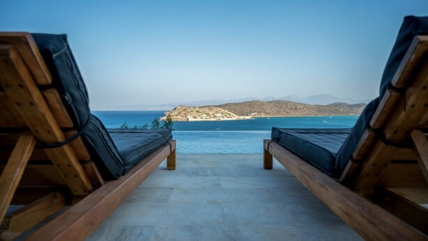 Two wooden lounge chairs facing a pool with a view of a beach and mountains in the distance. - Seafront Villa Astraiax, Divine Sea Views, Private Infinity Pool, Furnished. (Crete)