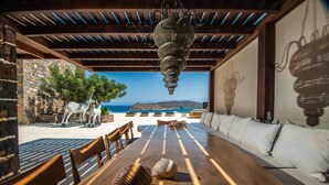 A wooden dining table with white cushions, a view of the sea, and a stone wall with a statue. - Seafront Villa Astraiax, Divine Sea Views, Private Infinity Pool, Furnished. (Crete)