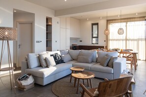 A modern living room with a sectional sofa, wooden side tables, and a dining area with a wooden table and chairs. - Villa Aurivis, Divine Sea Views, Private Infinity Pool, Furnished Terrace,. (Crete)