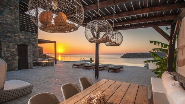 Un espace de restauration en plein air, avec une table en bois et des chaises en osier, offrant une vue sur le coucher de soleil sur la mer.
