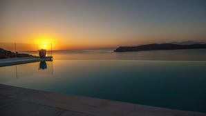 Une piscine à débordement aux eaux cristallines, offrant une vue imprenable sur la mer et les montagnes lointaines, le tout baigné par les couleurs du coucher de soleil.