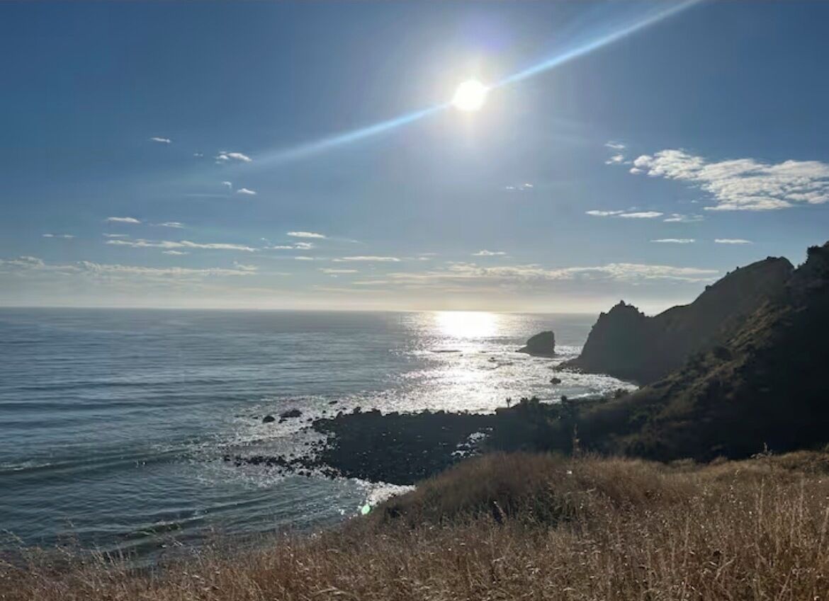 Paisaje costero con cielo despejado, el sol brillando y acantilados rocosos.