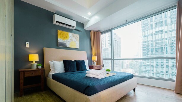 A modern hotel room with a large bed, a nightstand with a lamp, a wall-mounted air conditioner, and a large window offering a city view. - New! Comfortable 2BR in Central BGC w/ Fast WiFi (G3W2+RG Taguig, Metro Manila, Philippines)