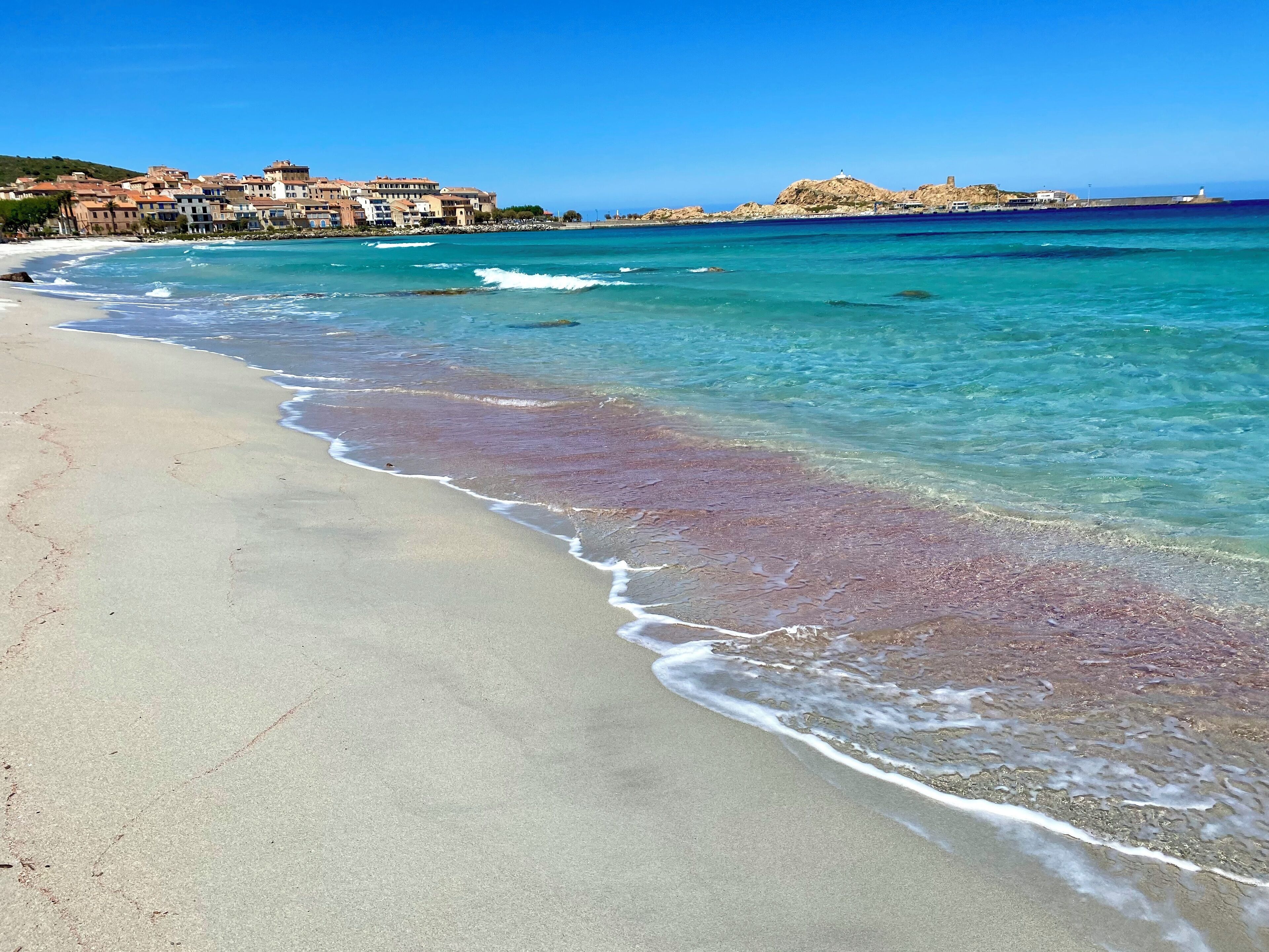 A beach with clear turquoise water, a sandy shore, and buildings in the background.