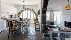 A spacious kitchen with white cabinets, a dining area with a large table and chairs, a stone fireplace, and a cozy seating area with a sofa and a coffee table. - Villa Elatheia, Prvate Pool and Garden, Furnished Terrace, BBQ facilities, Naxos (Naxos)