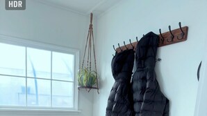 Una habitación con un banco de madera, un perchero con dos chaquetas abultadas negras, una planta colgante y una ventana con vista a un edificio.
