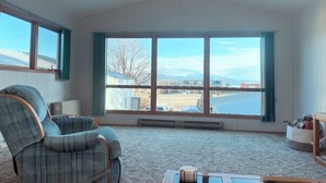 Amplia habitación con un gran ventanal que ofrece vistas a una masa de agua y montañas a la distancia. Hay un sillón escocés y una mesita con una taza y un platillo.