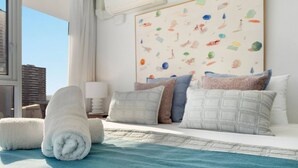 A hotel room with a bed, pillows, a rolled towel, a painting on the wall, and a window with curtains. - Beach Haven Getaway Broadbeach - WiFi, Parking (Broadbeach)