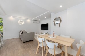 A modern, minimalist living space with a dining area, a sofa, and a TV mounted on the wall. - Salty`s at Burleigh - Central, Spacious, WiFi (Burleigh Heads)