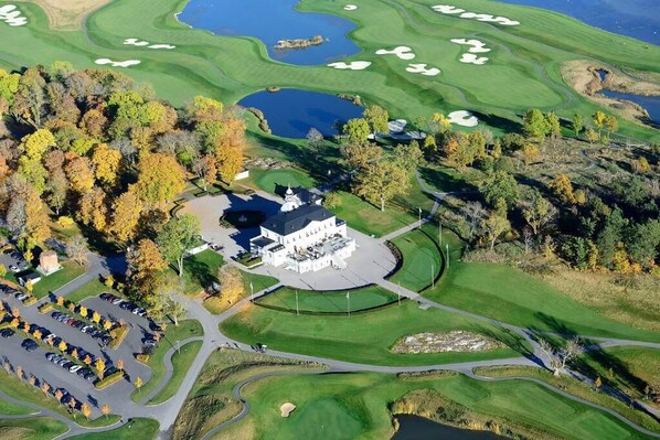 A golf course with a clubhouse, parking area, and surrounding trees.
