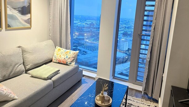 Ein modernes Wohnzimmer mit grauem Sofa, bunten Kissen, einem Couchtisch aus schwarzem Marmor und einem großen Fenster mit Blick auf die Stadt.