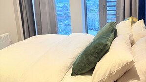 Ein Hotelzimmer mit einem großen Bett, vielen Kissen und Blick auf eine Stadtlandschaft durch die Fenster.
