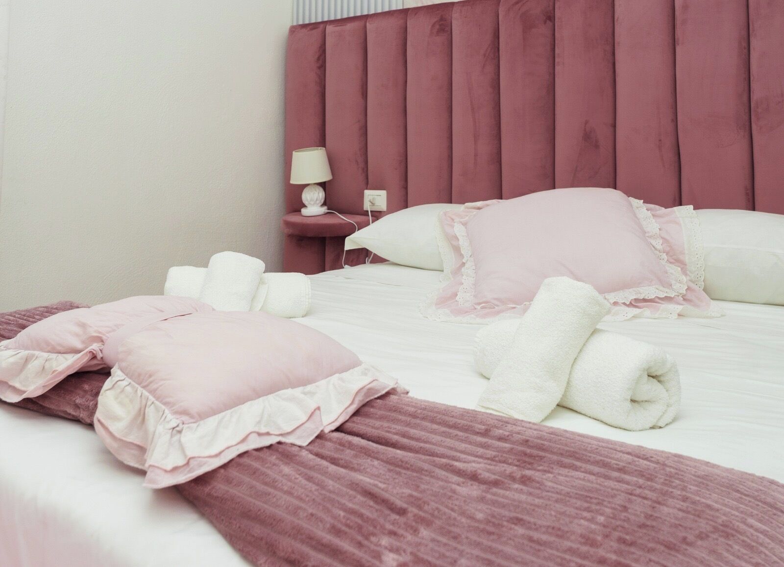 Una cama bien hecha con ropa de cama rosa y blanca, un cabecero rojo y una mesita de noche con una lámpara y un objeto decorativo pequeño.